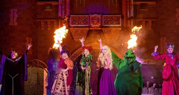 Disney villains perform on stage with dramatic gestures, flanked by flames, in front of a castle backdrop during a nighttime theatrical show.