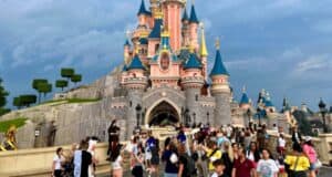A bustling scene of visitors at Disneyland Paris in front of the iconic Sleeping Beauty Castle, with its pink and blue spires under a cloudy sky.