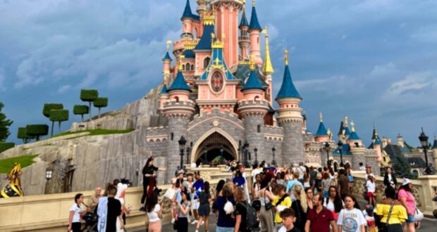 A bustling scene of visitors at Disneyland Paris in front of the iconic Sleeping Beauty Castle, with its pink and blue spires under a cloudy sky.