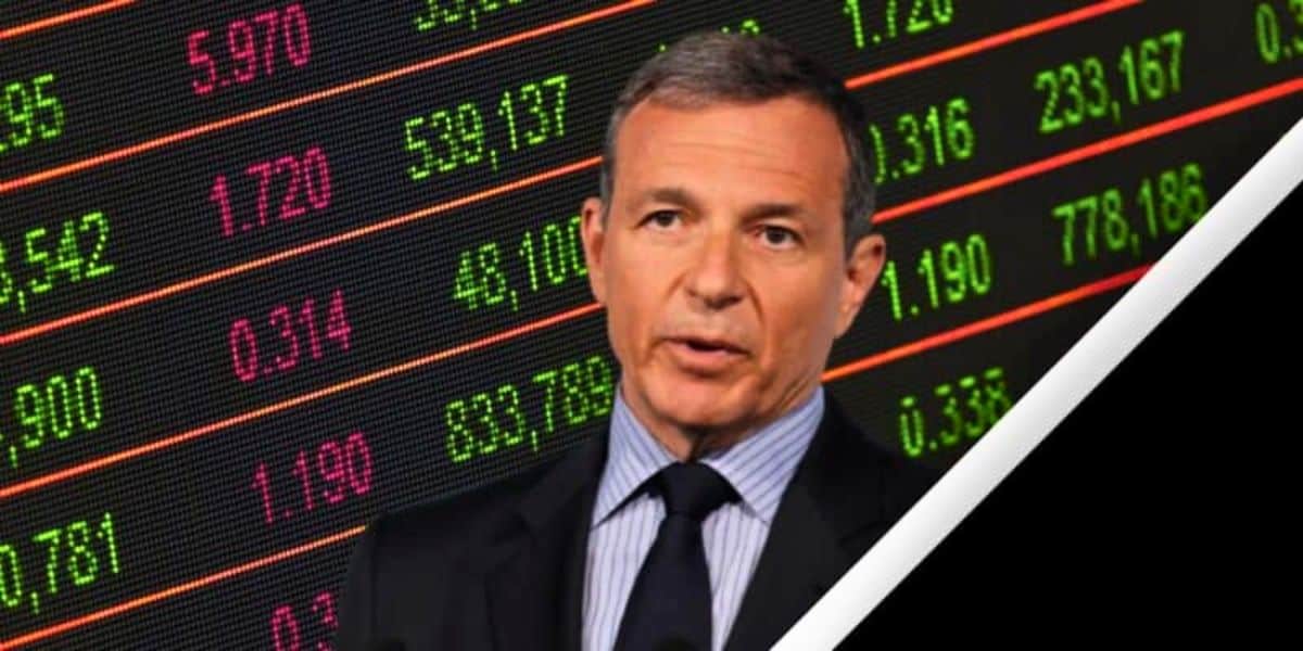 A man in a suit and striped tie speaks in front of a digital stock market display board showing various numbers and figures in red and green, including Disney Stock. The image has a diagonal white and black border on the right side.
