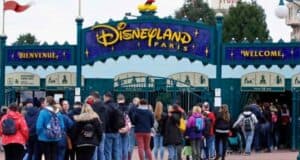 A crowd of people waits in line to enter Disneyland Paris. The main entrance features a large sign with "Disneyland Paris" in colorful letters, and a smaller "Welcome" sign underneath. Many visitors carry backpacks and wear winter clothing.