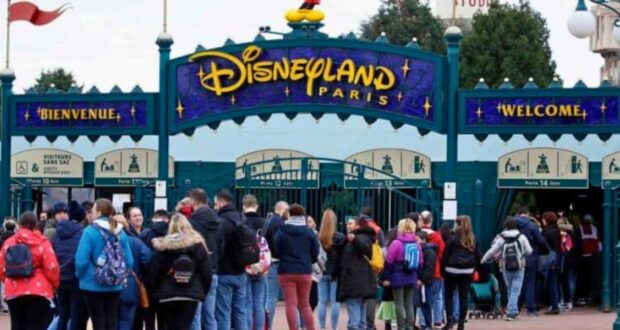A crowd of people waits in line to enter Disneyland Paris. The main entrance features a large sign with "Disneyland Paris" in colorful letters, and a smaller "Welcome" sign underneath. Many visitors carry backpacks and wear winter clothing.