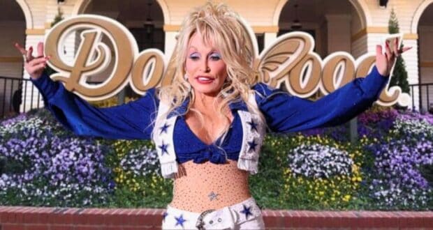 A person with long blonde hair and a sparkly blue and white outfit standing in front of the Dollywood sign, channeling their inner Dolly Parton. They are smiling with their arms raised. The background includes colorful flowers and part of a building.