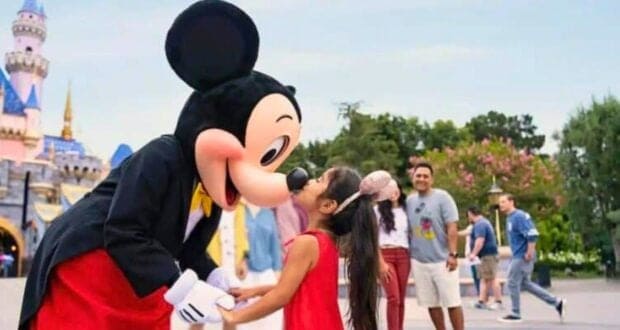 A girl in a red dress happily kisses Mickey Mouse on the nose at Disney, with other visitors and the castle blurred in the background.