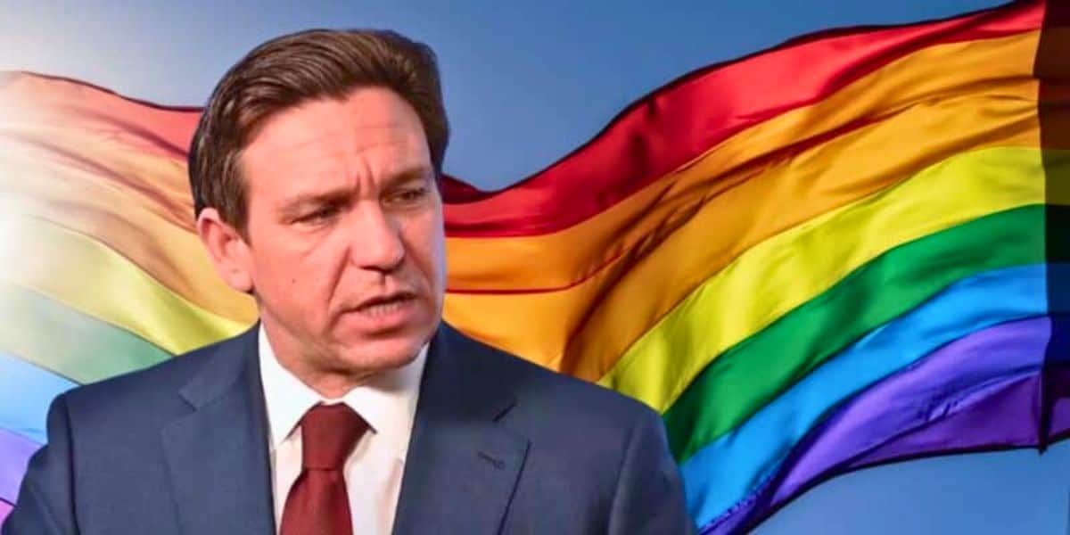 A person in a suit and tie is shown in front of a large rainbow flag. The flag's colors are vibrant, waving against a clear sky. The person appears to be making an expression as they look slightly to the right.
