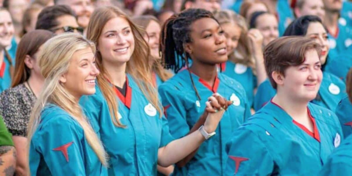 A diverse group of people, mostly young adults, are standing closely together, smiling, and looking ahead. They are all wearing teal uniforms with badges. Some are holding hands, creating a sense of unity and celebration. The background shows more people in similar attire.