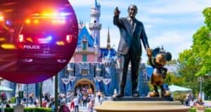 A statue of Walt Disney holding Mickey Mouse's hand stands in front of Sleeping Beauty Castle at Disneyland. Inserted in the image is a photo of a police car with flashing lights at night. Visitors walk around the statue and castle in the background.