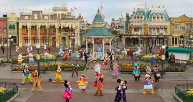 A lively scene at what appears to be a Disney theme park features a variety of costumed characters, including Mickey Mouse, Minnie Mouse, and Donald Duck, performing in front of colorful, ornate buildings and a gazebo on Main Street.