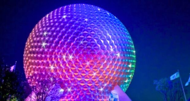 A large geodesic sphere, known as Spaceship Earth, is illuminated in vibrant rainbow colors against the night sky. Flags can be seen in the bottom right near trees silhouetted by the glow of the lights.