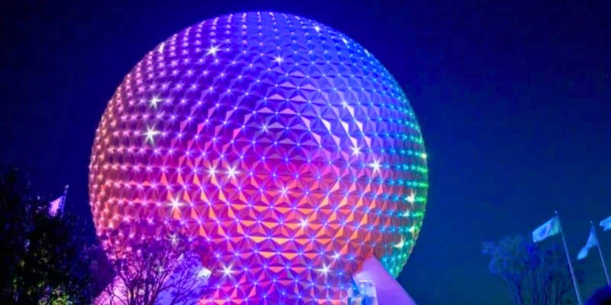 A large geodesic sphere, known as Spaceship Earth, is illuminated in vibrant rainbow colors against the night sky. Flags can be seen in the bottom right near trees silhouetted by the glow of the lights.