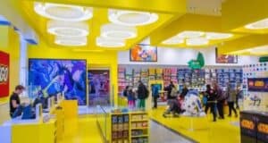 A vibrant LEGO store with bright yellow walls and ceiling invites enthusiasts to explore. Shoppers admire displays featuring various sets, including a standout R2-D2 model in the Star Wars section. Fans gather eagerly around diverse product areas, sharing their passion for the LEGO universe.
