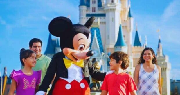 A family of four beams with joy standing next to a cheerful Mickey Mouse at a magical theme park. Behind them, a majestic castle with blue spires rises against the clear sky, capturing their excitement and happiness.