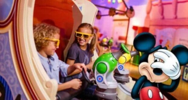 Two children seated in a Disneyland ride vehicle with 3D glasses, smiling and holding blasters. A whimsical backdrop and colorful lighting enhance the amusement park setting. Meanwhile, a life-sized Mickey Mouse figure stands nearby, playfully covering his face with one hand, perhaps bashfully avoiding the viral hack that's been criticized for destroying Disney property.