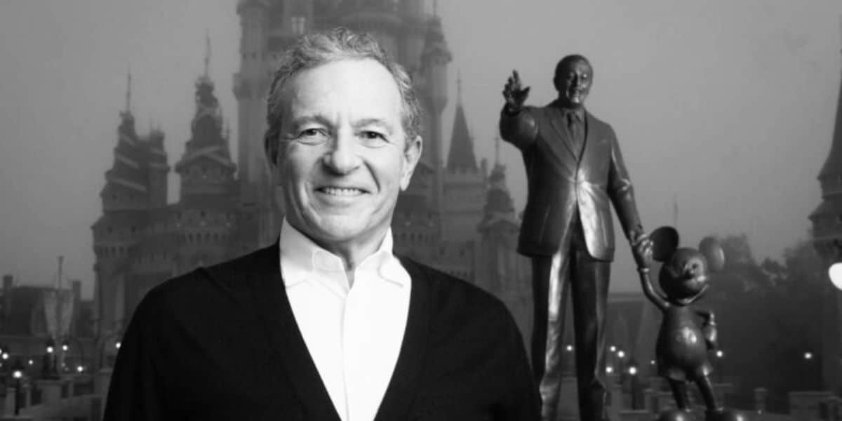 A man smiles broadly while standing in front of a castle and a statue of a man holding hands with a mouse character, iconic symbols of the Disney Company. The image is in black and white, and the atmosphere appears slightly foggy.