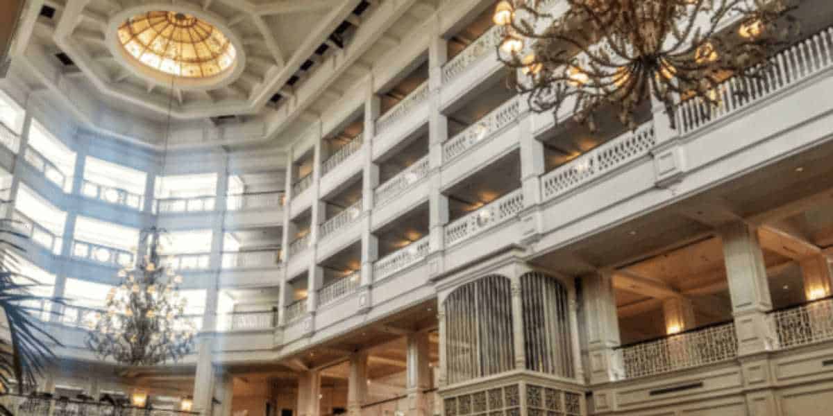 An opulent hotel atrium with a multi-story layout, featuring balconies lined with white railings. A grand chandelier hangs centrally, and sunlight streams from a large, ornate dome at the top, illuminating the space with a warm glow.