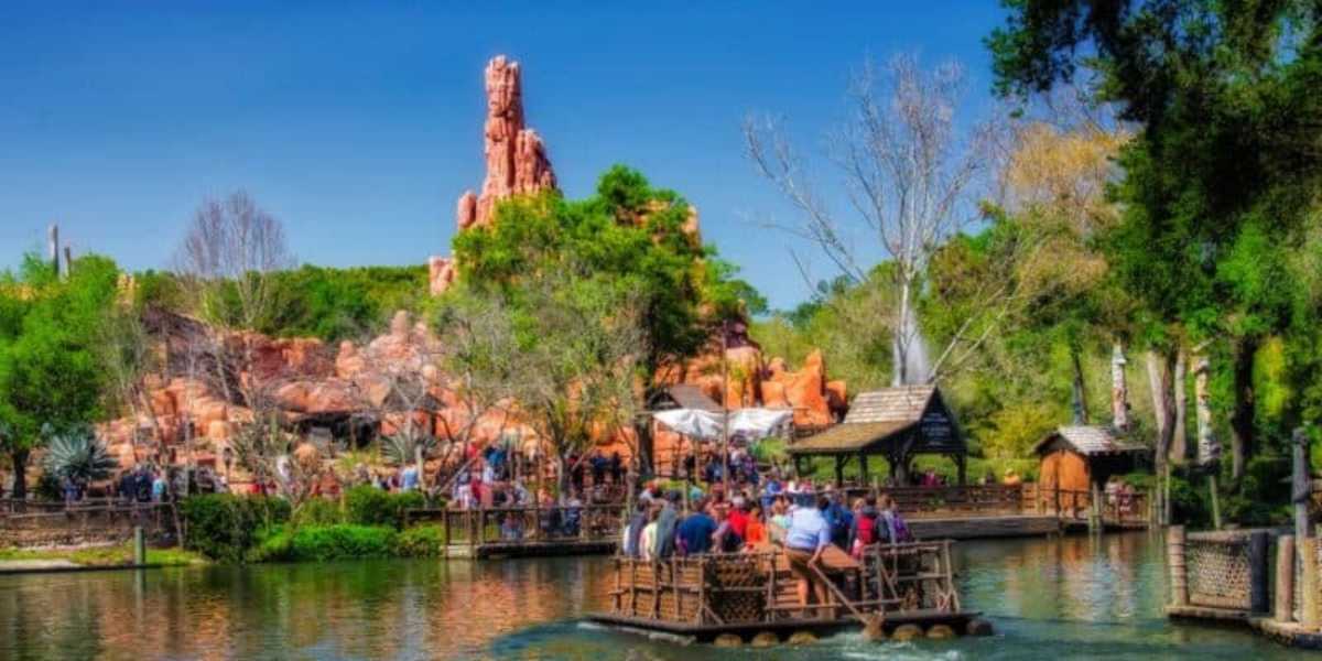 A lively amusement park scene featuring a ride over a peaceful body of water, with people on a raft in the foreground. In the background, visitors walk near a rustic structure and a rocky, mountainous attraction surrounded by lush greenery. The sky is clear and blue.