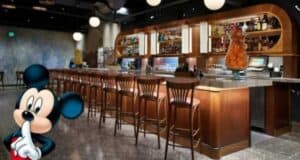 A modern bar with wooden chairs and a marble countertop is shown. Various bottles and glasses are displayed on shelves behind the bar. A figure of Mickey Mouse is in the foreground, holding a finger to his lips as if signaling to be quiet, capturing the secretive vibe of Enzo's Hideaway.