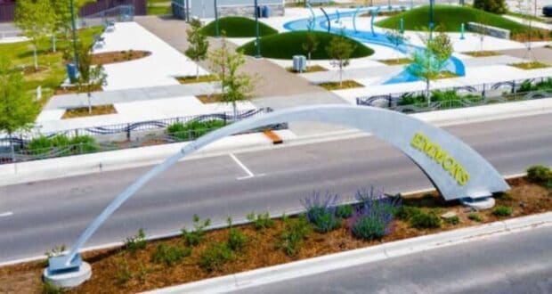 A modern public park featuring a large arched structure with the word "EMMONS" on a street median. The park has curved pathways, grassy mounds, young trees, and landscaped areas with purple flowers. The scene is vibrant and well-maintained.