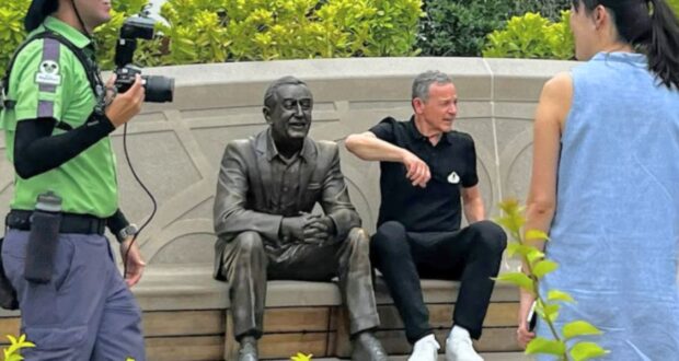 A man in a black shirt sits on a bench beside a bronze statue of Walt Disney. The man points or gestures in conversation with a woman in a sleeveless blue dress facing him. Another person in a green shirt and cap holds a camera, appearing to capture the meeting.