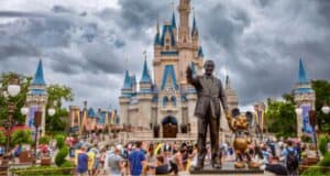 A vibrant scene at a theme park featuring a large castle with blue turrets under a cloudy sky. In the foreground, a statue of a person holding hands with a cartoon mouse overlooks a bustling crowd of visitors. Lush greenery and flags decorate the surroundings.