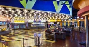 An empty and brightly lit cinema lobby features patterned tile flooring, multiple concession stands with illuminated signs advertising meals, and organized queues. The colorful ceiling with geometric shapes enhances the lively atmosphere, while the seating area is visible in the background.