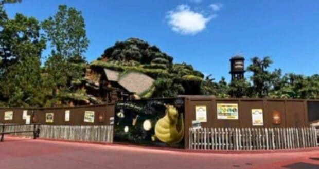 A large construction wall surrounds Tiana's Bayou Adventure, a mountain with greenery and a rocky hill reminiscent of Splash Mountain visible in the background.