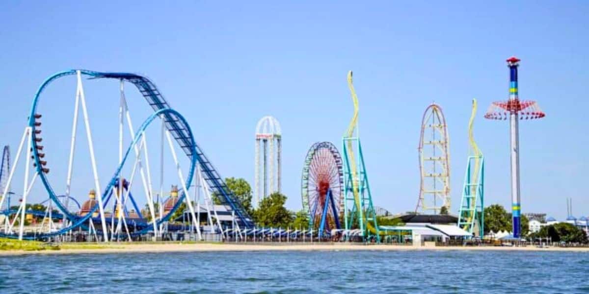 Image of Cedar Point amusement park with large roller coasters, a Ferris wheel, and a tall drop tower. The park is situated beside a body of water, with clear blue skies overhead. The vibrant rides and structures stand out against the green trees and open space.