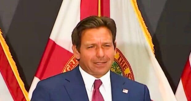 Ron DeSantis is standing in front of a backdrop featuring red and white flags. He has short brown hair and is speaking or making a gesture with his right hand.