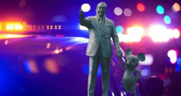 A statue of a man in a suit holding hands with a cartoon mouse is in the foreground. Behind them are blurred police lights with red and blue hues, illuminating the scene.
