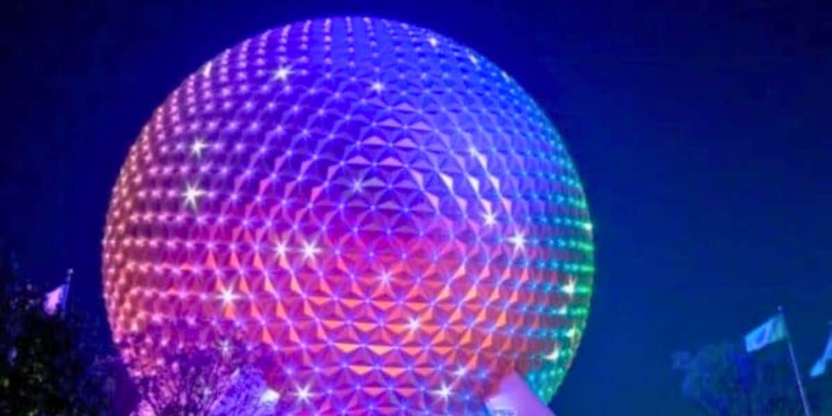 A large geodesic sphere, reminiscent of Remo Saraceni's imaginative designs, is illuminated with a gradient of colors from purple to orange, creating a vibrant display against the dark night sky. The surface is composed of triangular panels with twinkling lights. Flags and trees are partially visible nearby.