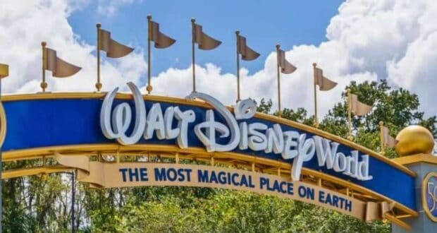 An entrance sign to walt disney world, featuring a large blue banner with the park's name in stylized white lettering, flanked by flags, under a clear blue sky with fluffy clouds.