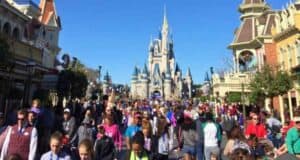 A bustling crowd fills the street leading to a whimsical castle at a theme park. The clear blue sky and colorful buildings create a vibrant atmosphere as visitors explore the attractions and shops. The iconic castle stands tall in the background.