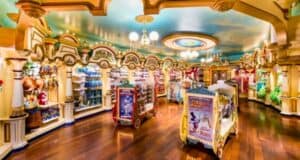A brightly lit, colorful toy store with wooden floors and ornate architecture. Shelves are lined with a variety of toys, including stuffed animals and action figures. Display stands in the center feature themed Disney merchandise. A painted sky ceiling adds to the whimsical atmosphere.