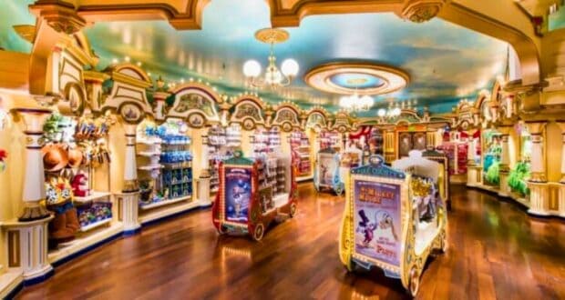 A brightly lit, colorful toy store with wooden floors and ornate architecture. Shelves are lined with a variety of toys, including stuffed animals and action figures. Display stands in the center feature themed Disney merchandise. A painted sky ceiling adds to the whimsical atmosphere.