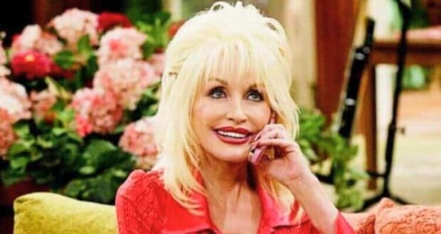 Dolly Parton with blond hair and a bright smile is sitting on a couch, holding a pink phone to her ear. Behind her, there are colorful flowers and green plants in a cozy indoor setting.