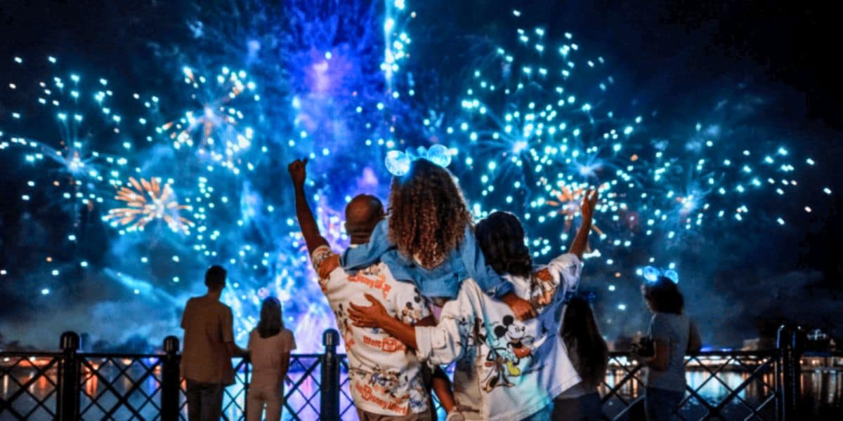A group of people watches a vibrant fireworks display at night, with one person wearing glowing mickey mouse ears. they are standing by a railing, illuminated by the colorful bursts in the sky.