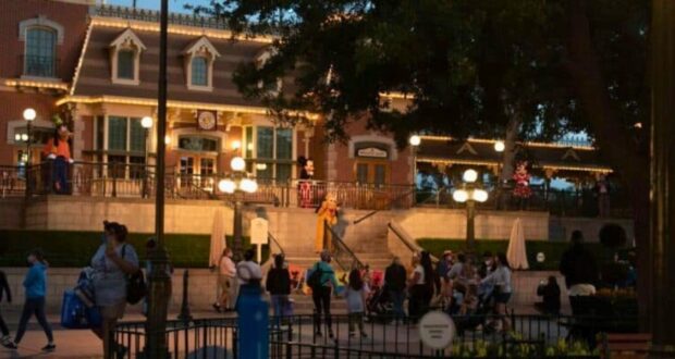 A lively evening scene at a Disney theme park entrance, featuring beautifully lit buildings adorned with lights. Characters in costumes interact with guests, some of whom are wearing masks. The area is filled with people strolling and taking in the festive atmosphere.