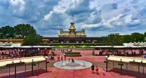 View of the entrance to Disney World, featuring a railway station in the background, bustling with visitors under a cloudy sky.