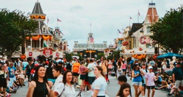 A bustling theme park scene unfolds along Main Street America, with a crowd of people walking past buildings adorned with festive bunting and American flags. Some visitors rest on the ground, while a cloudy sky looms overhead.