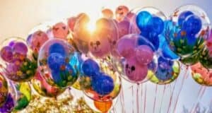 A cluster of colorful balloons floats against a bright, sunny sky. The balloons are inside clear outer balloons with vibrant designs, including flowers, butterflies, and cartoon faces, creating a whimsical and joyful scene. In the background, you can slightly see trees as a dad refuses to take his daughter to Disney.