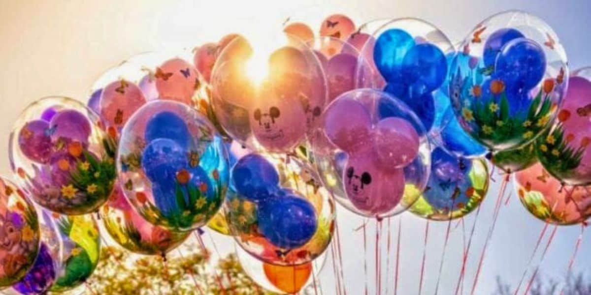 A cluster of colorful balloons floats against a bright, sunny sky. The balloons are inside clear outer balloons with vibrant designs, including flowers, butterflies, and cartoon faces, creating a whimsical and joyful scene. In the background, you can slightly see trees as a dad refuses to take his daughter to Disney.