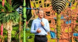 A person in a light blue shirt stands outdoors, holding a microphone and speaking. The background features colorful, eclectic decorations and greenery, creating a vibrant atmosphere.