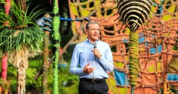 A person in a light blue shirt stands outdoors, holding a microphone and speaking. The background features colorful, eclectic decorations and greenery, creating a vibrant atmosphere.