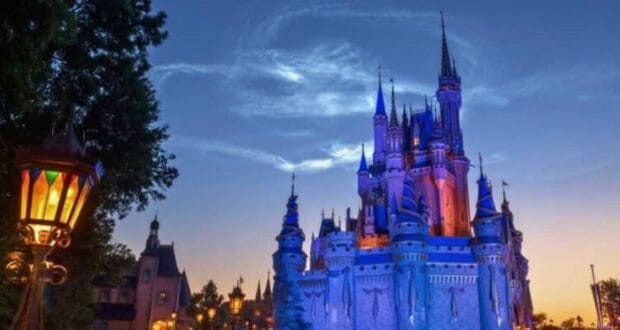 A fantasy castle illuminated in blue and purple lights with towering spires stands against a twilight sky. Nearby, a colorful medieval-style lantern glows, and shadowy trees frame the scene. It's a sight so magical that even a Drunk Disney World Guest might find themselves momentarily arrested by its beauty.