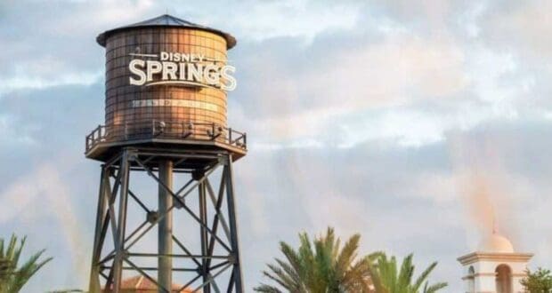 A tall water tower with "Disney Springs" written on it stands against a partly cloudy sky. Palm trees and a white building with a bell tower are visible in the background.