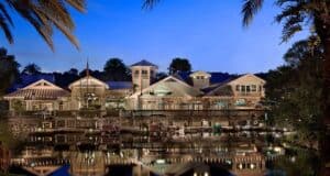 A resort building with a waterfront view, illuminated at twilight. The structure features a combination of modern and rustic architectural styles, surrounded by palm trees. The calm water in front reflects the lights and trees, creating a picturesque scene reminiscent of the Gurgling Suitcase at Disney's Old Key West Resort.