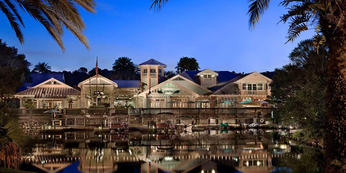 A resort building with a waterfront view, illuminated at twilight. The structure features a combination of modern and rustic architectural styles, surrounded by palm trees. The calm water in front reflects the lights and trees, creating a picturesque scene reminiscent of the Gurgling Suitcase at Disney's Old Key West Resort.