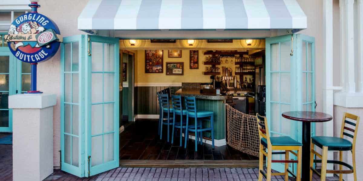 A small bar with turquoise folding doors open, revealing a cozy interior with stools at the counter and framed photos on the walls. Outside, a round table with colorful chairs sits under a striped awning. A sign reads "Gurgling Suitcase" at Disney's Old Key West Resort.