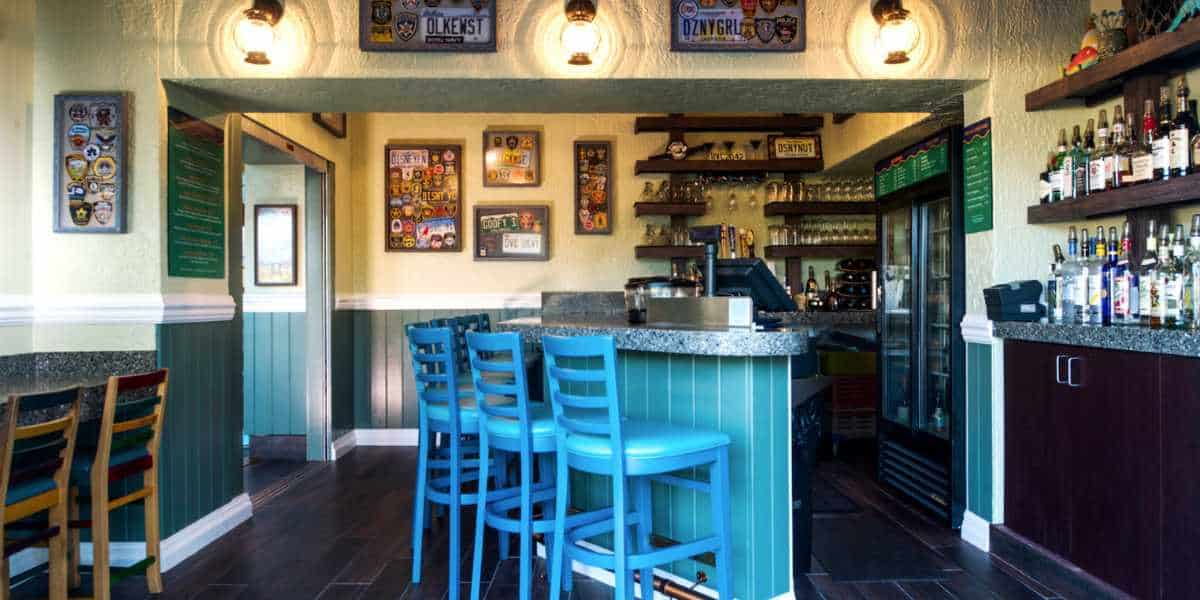 A cozy bar interior at the Gurgling Suitcase at Disney's Old Key West Resort features a row of blue bar stools along a counter. The walls are decorated with various beer logos and memorabilia. Shelves behind the counter display an assortment of bottles. Warm lighting creates a welcoming atmosphere.