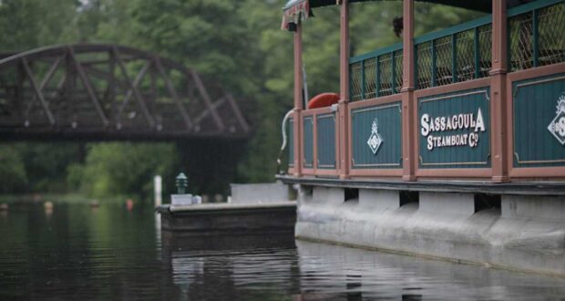 A Disney boat with green and brown colors, displaying the name "Sassagoula Steamboat Co.," is moored on a tranquil river with a trestle bridge partially visible in the background and surrounded by lush green trees, providing charming transportation through scenic landscapes.